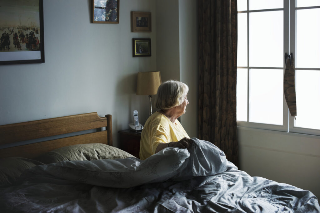 Senior woman sitting up in bed, looking out the window on a grey day.