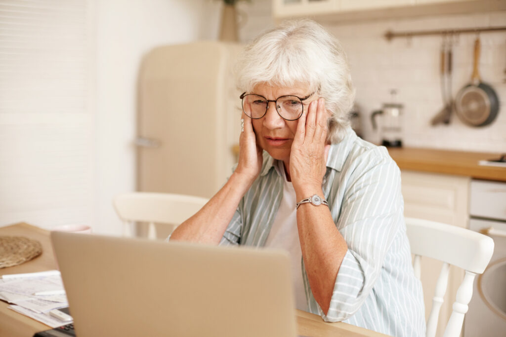 Lonely upset retired woman with gray hair having frustrated stressed look, touching head, facing tech problems, using laptop
